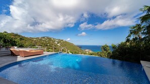 Une piscine avec des chaises longues, une terrasse en bois et une vue sur l’océan et les collines.