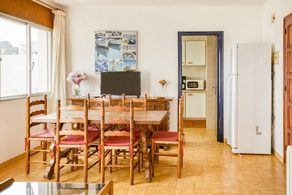 A dining area with a wooden table and chairs, a flat-screen TV, and a kitchen with a microwave and refrigerator.