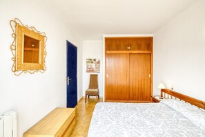 A bedroom with a bed, a wooden wardrobe, a chair, a small table, and a wall-mounted mirror.