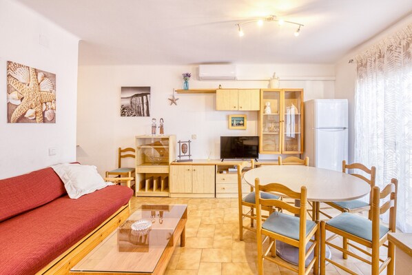A compact living space with a sofa, dining table, chairs, a TV, kitchen cabinets, and a refrigerator.