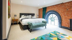 A bedroom with a large bed, a bench, a nightstand, a lamp, and a view through a window with a brick wall. - Apartment 2 The Old Chapel apartments, in the heart of Sherwood Forest (Edwinstowe, Nottinghamshire)