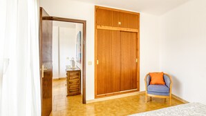 A bedroom with a wooden wardrobe, a bed with patterned bedding, a chair with an orange cushion, and a wooden dresser.