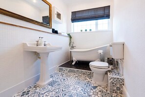 A bathroom with a white pedestal sink, a freestanding bathtub, and a toilet. The floor has patterned tiles, and there is a window with a dark valance. - Riverside Rest - quirky split-level 3-bed cottage (Ollerton, Nottinghamshire)