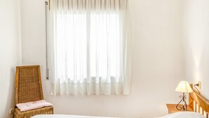 Ein Schlafzimmer mit einem Bett, einem Korbstuhl mit Kissen, einem Nachttisch mit Lampe und einem Fenster mit durchsichtigen Vorhängen.