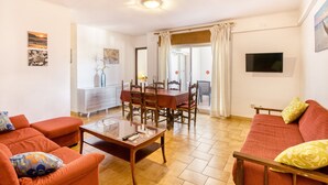 Ein Wohnzimmer mit einer Couch, einem Couchtisch, einem Esstisch mit Stühlen, einem Fernseher und einer Glasschiebetür, die auf einen Balkon führt.