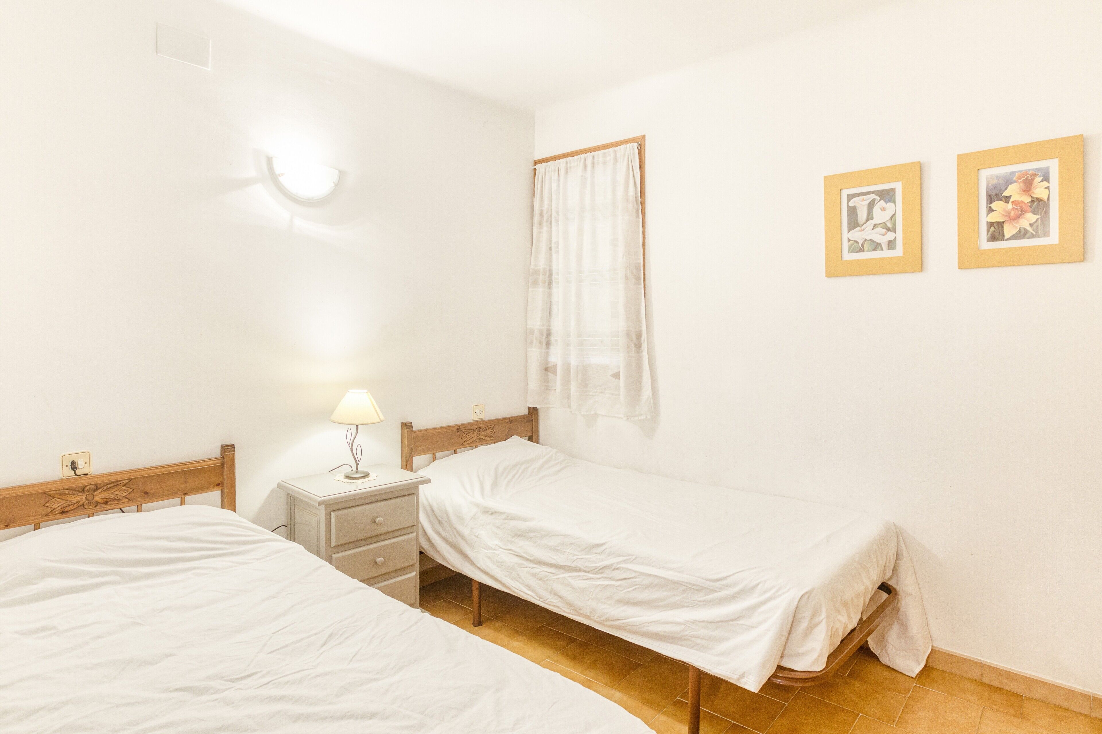 A bedroom with two single beds, a nightstand with a lamp, a window with a sheer curtain, and two framed pictures on the wall.