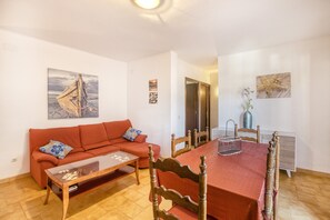 Ein Wohnzimmer mit einem roten Sofa, einem Glastisch, einem Holzesstisch mit roter Tischdecke und einem Gemälde eines Bootes an der Wand.