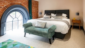Une chambre avec un grand lit, un fauteuil vert, un mur en briques et une fenêtre.
