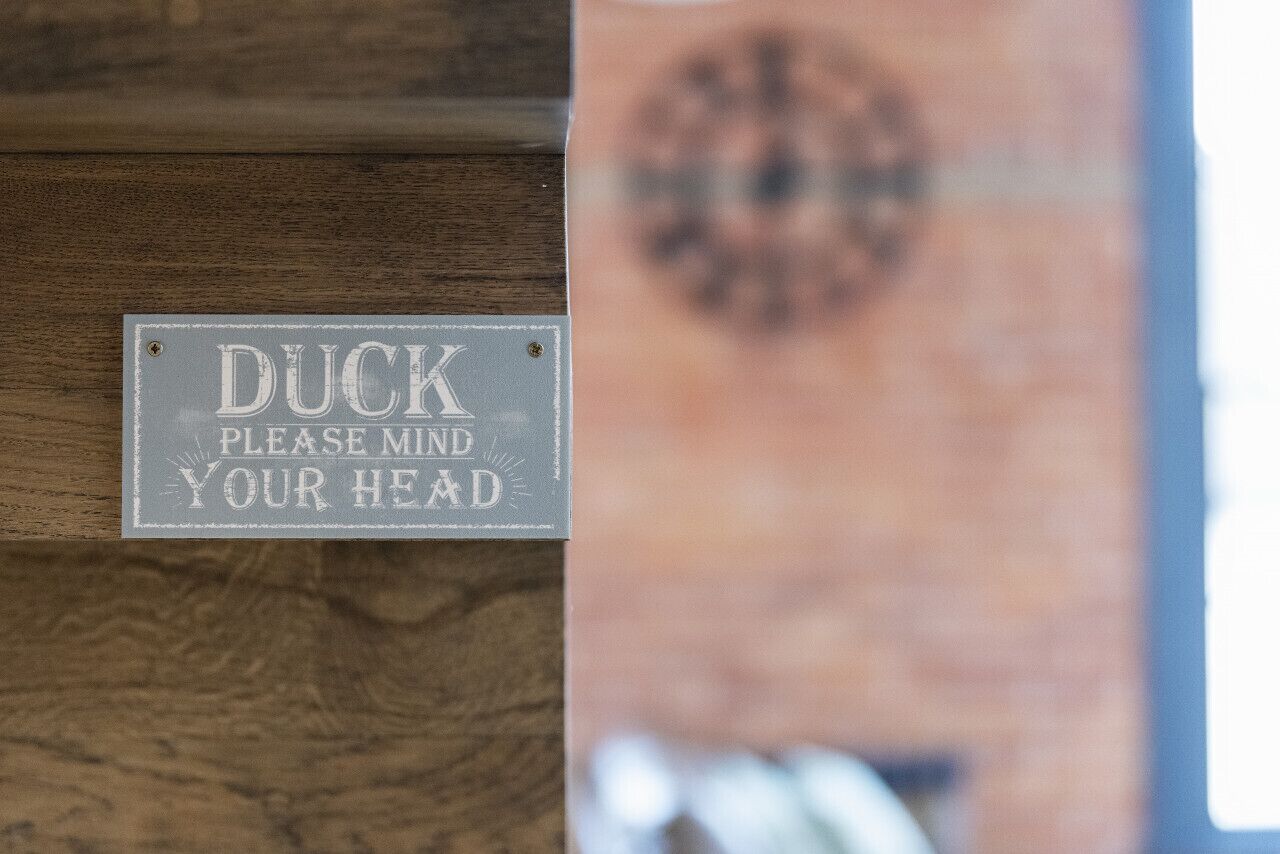 A sign on a wooden surface that reads "DUCK PLEASE MIND YOUR HEAD".