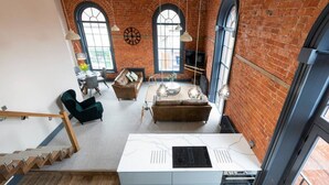 Un loft spacieux avec des murs en briques apparentes, de grandes fenêtres, une cuisine moderne dotée d’une cuisinière intégrée et un coin salon confortable avec un canapé et des fauteuils.