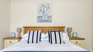 A neatly made bed with a blue quilt, two bedside tables with lamps, and a painting of a vase with flowers on the wall. - Ropemaker's Cottage, · Ropemaker's Cottage, cosy cottage retreat (Ollerton, Nottinghamshire)