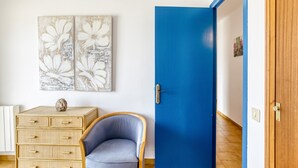 Una habitación con una puerta azul, un aparador de mimbre, un sillón azul y dos cuadros florales.