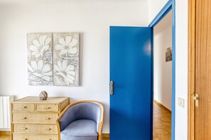 A room with a blue door, a wicker chest of drawers, a blue armchair, and two floral wall art pieces.