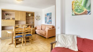 Una sala de estar con un sofá, una mesa de comedor con sillas, una zona de cocina con armarios y una pared con una pintura de frutas.
