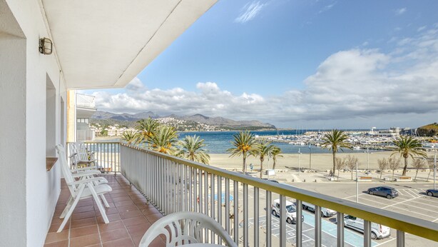 Un balcón con sillas y una mesa blancos, con vista a una playa con palmeras y un puerto deportivo.