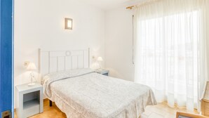 Un dormitorio con cama, mesitas de noche, una lámpara de pared y una ventana con cortinas traslúcidas.