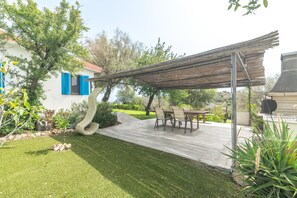 A patio with a table and chairs, surrounded by a garden and a white building with blue shutters.