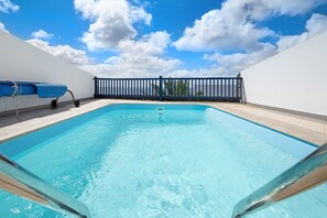 En klar, blå swimmingpool med en metalstige, en blå liggestol og et sort hegn.