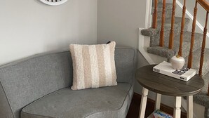 Une pièce d’angle avec un canapé gris, un miroir rond, une petite table ronde sur laquelle se trouvent un livre et un vase, et un escalier en bois.