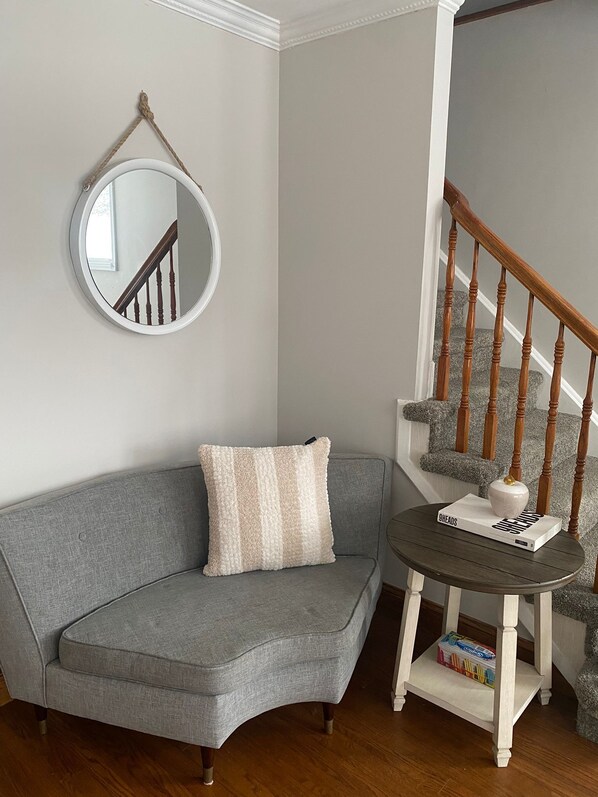A corner room with a grey sofa, a round mirror, a small round table with a book and a vase, and a wooden staircase.