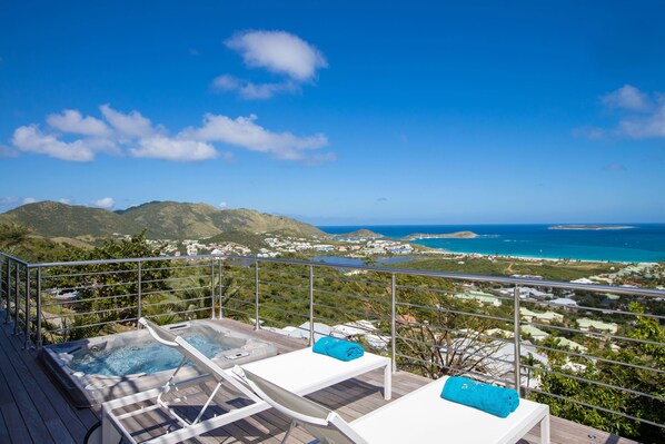 Un balcón con dos jacuzzis y sillones de descanso, con vistas a un paisaje costero con montañas y al mar.