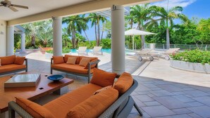 A modern outdoor lounge area with wicker furniture, a pool, and a view of palm trees and a clear sky. - Les Palmiers Bleus Luxury villa (Les Terres Basses)