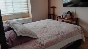 Een slaapkamer met een bed, een aan de muur bevestigde televisie, een raam met zonwering en een nachtkastje met een lamp.