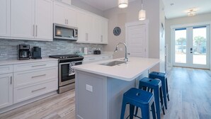A modern kitchen with white cabinets, a stainless steel stove, a microwave, and a sink. There are three blue bar stools and a clock on the wall. - New!! Steps To Beach, Gulf Views, Private Heated Pool (Panama City Beach)
