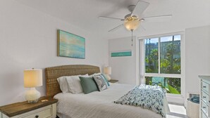 Una camera da letto con un letto, comodini, una ventola a soffitto, una finestra con vista sul verde e un cartello appeso al muro.