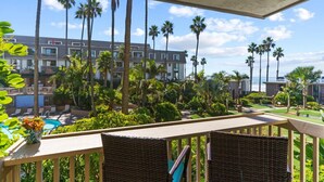 Un balcone con sedie in vimini, una piscina e vista su un hotel con palme e cielo sereno.