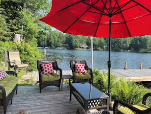 A wooden deck with wicker furniture and a red umbrella, overlooking a lake with boats and houses in the background.