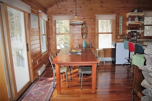 A wooden cabin interior with a dining table, chairs, a kitchen area, and a stone fireplace.
