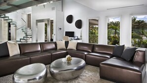 Un salon moderne avec un canapé d’angle, deux petites tables rondes et une grande fenêtre donnant sur un paysage verdoyant.
