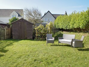 Een achtertuin met een houten tuinhuisje, twee rieten stoelen en een haag.