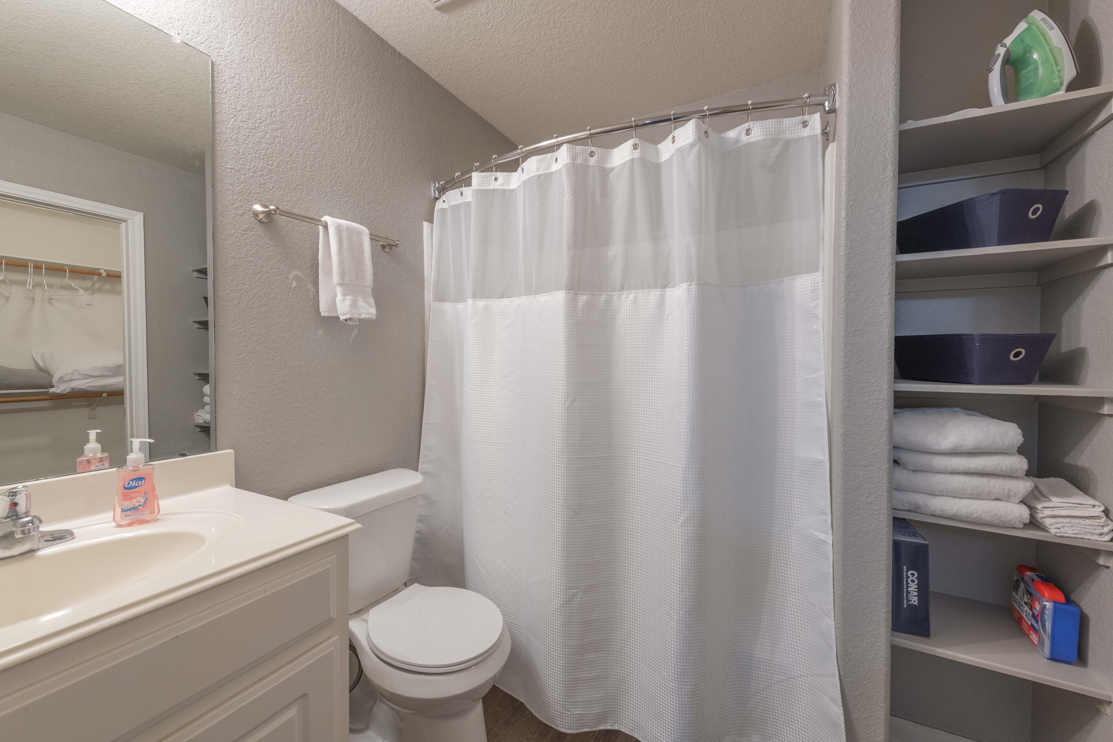 A bathroom with a white sink, toilet, and shower curtain. There are shelves with folded towels and storage boxes.