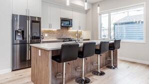 A modern kitchen with stainless steel appliances, a central island with bar stools, and a large window providing natural light. - Executive home, Rooftop Deck, close to Foothills Med Ctr, Kensington Plaza & DT (Calgary)