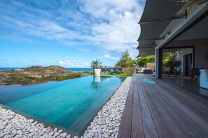 An infinity pool with a view of the ocean, a wooden deck, and a covered outdoor seating area.