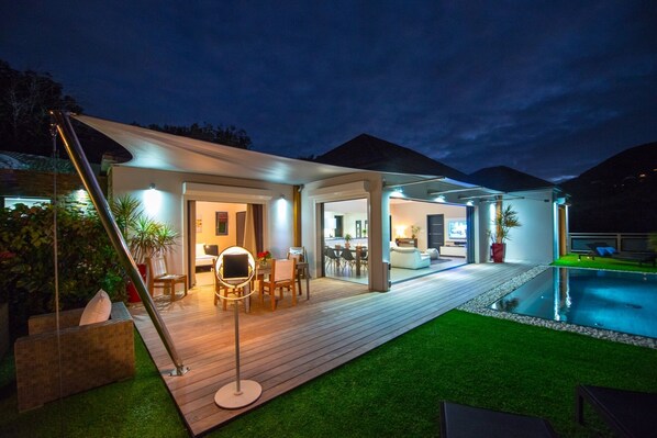 A modern outdoor living area with a pool, wooden deck, and covered seating area.