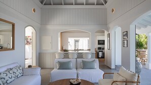 A spacious living area with a sofa, chairs, and a coffee table. There is a kitchen area with a sink and cabinets, and a dining area with chairs. The room has a high ceiling with wooden beams and large windows. - Beautiful Bijou Luxury villa (Flamands)
