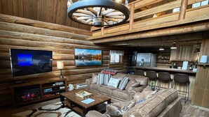 A log cabin interior with a leather sofa, a mounted flat-screen TV, a chandelier, and a kitchen area. - Rustic MT Cabin (Kalispell)