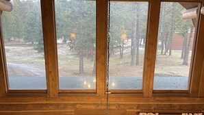 A wooden cabin interior with a central wooden chandelier, large windows, and a view of the surrounding forest. - Rustic MT Cabin (Kalispell)