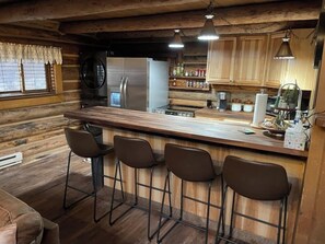 A rustic kitchen with wooden walls, a stainless steel refrigerator, a wooden bar counter with bar stools, and hanging pendant lights.