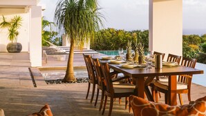 Un espacio de comedor al aire libre con una mesa lista para seis personas, rodeada de sillas, cerca de una piscina y una palmera.