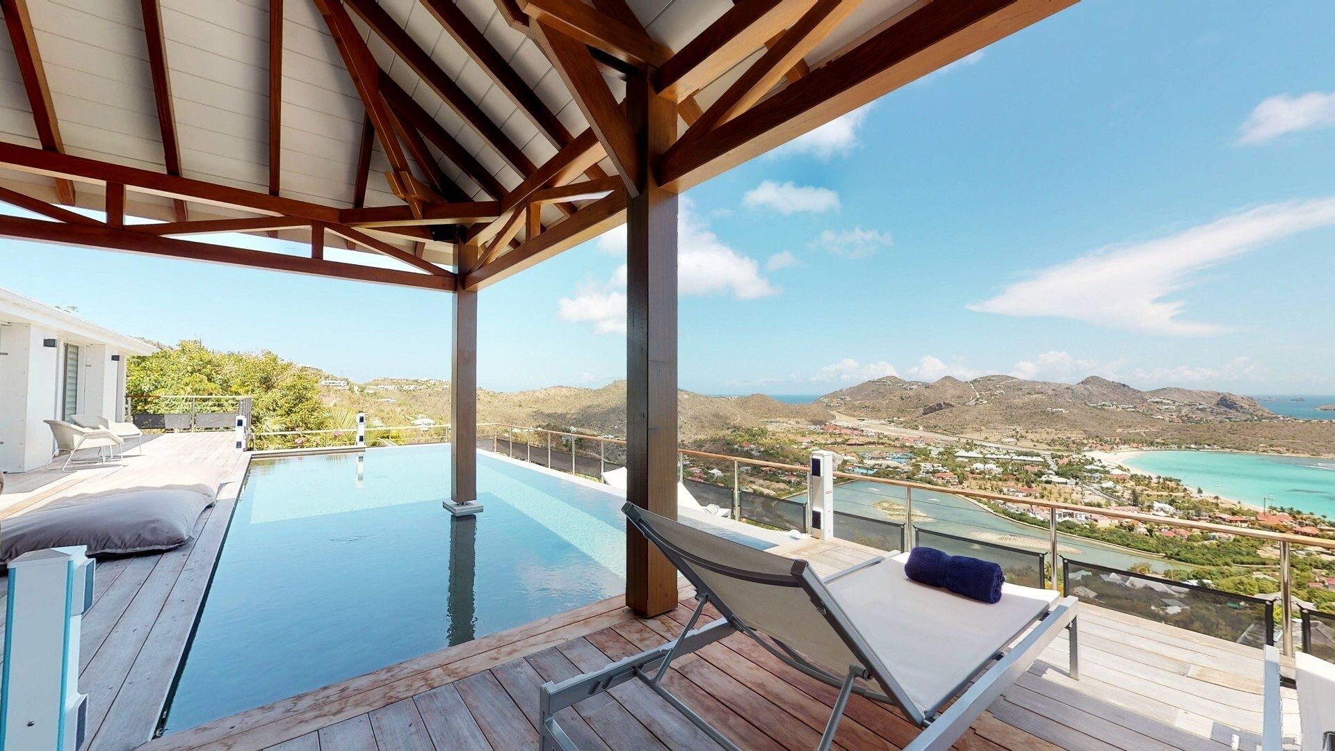 Une piscine à débordement avec une terrasse en bois, une chaise longue et une vue sur un paysage côtier avec des montagnes.