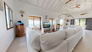 A spacious living area with a large white sofa, a wooden cabinet, and a television on a white stand. There are ceiling fans, a large window with a view, and a table with chairs. - W View (1/4 Rooms) Luxury villa (Saint Jean)