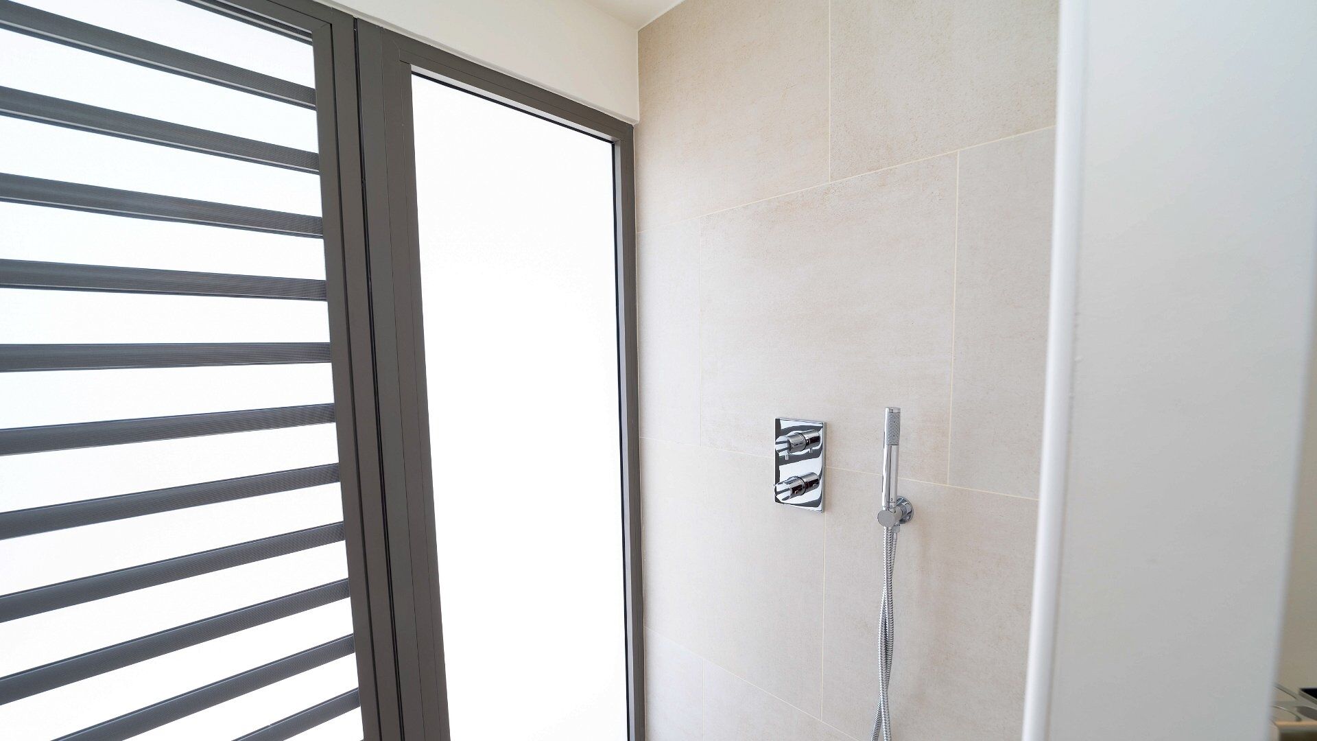 A bathroom with a shower, a window with blinds, and a tiled wall.