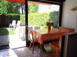 An open patio with a table and chairs, a potted plant, and a view of a garden and a building in the background.