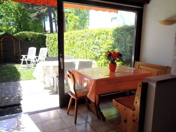 An open patio with a table and chairs, a potted plant, and a view of a garden and a building in the background.