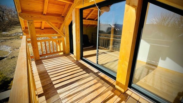 Eine Holzterrasse mit großen Fenstern, einem Hollerleger und freiem Blick auf die umliegende Landschaft.