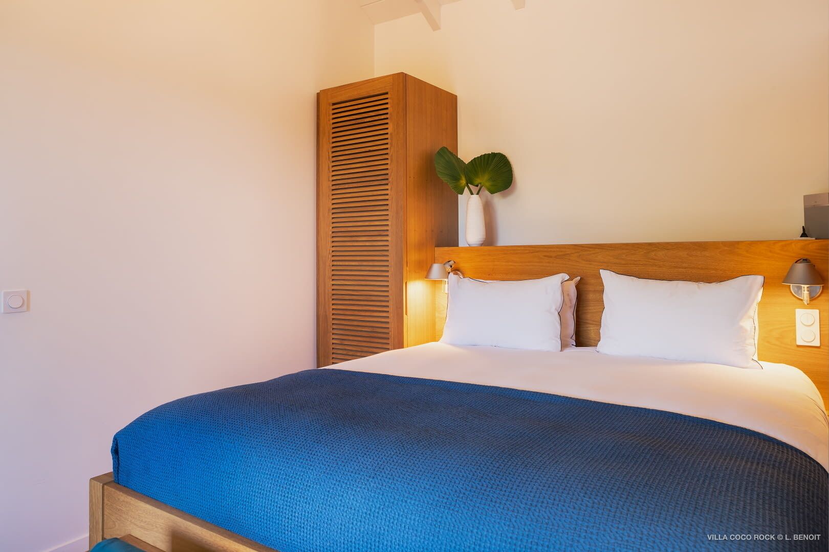 Habitació d'hotel amb un capçal de llit de fusta, un llit amb roba de llit blanca, una manta blava i una planta en test amb la taula de nit.